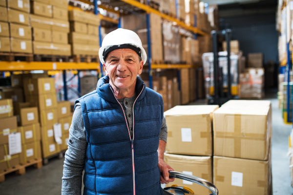 Senior male warehouse worker or a supervisor with white helmet pulling a pallet truck with boxes.