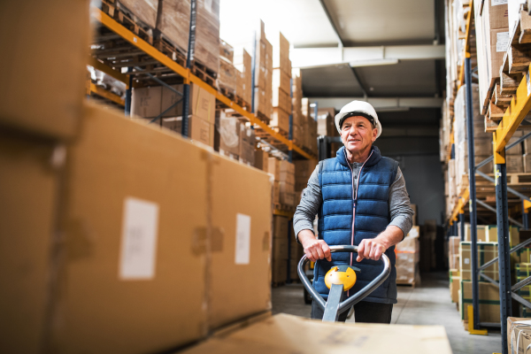 Senior male warehouse worker pulling a pallet truck with boxes.