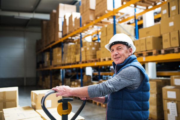 Senior male warehouse worker or a supervisor with white helmet pulling a pallet truck with boxes.