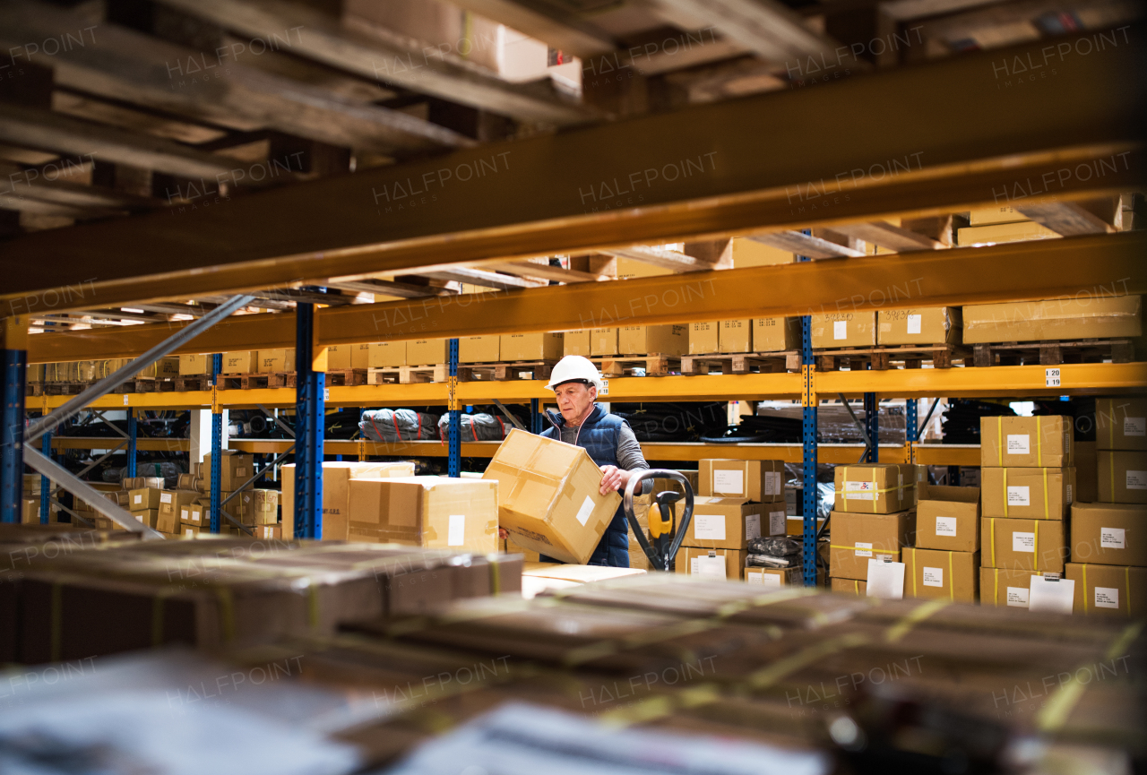 Senior male warehouse worker or a supervisor with white helmet loading or unloading a pallet truck with boxes.