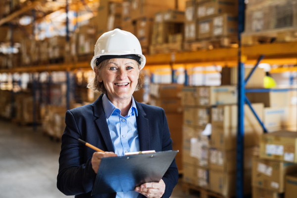 Senior woman warehouse manager or supervisor with a white helmet and clipboard, controlling stock.