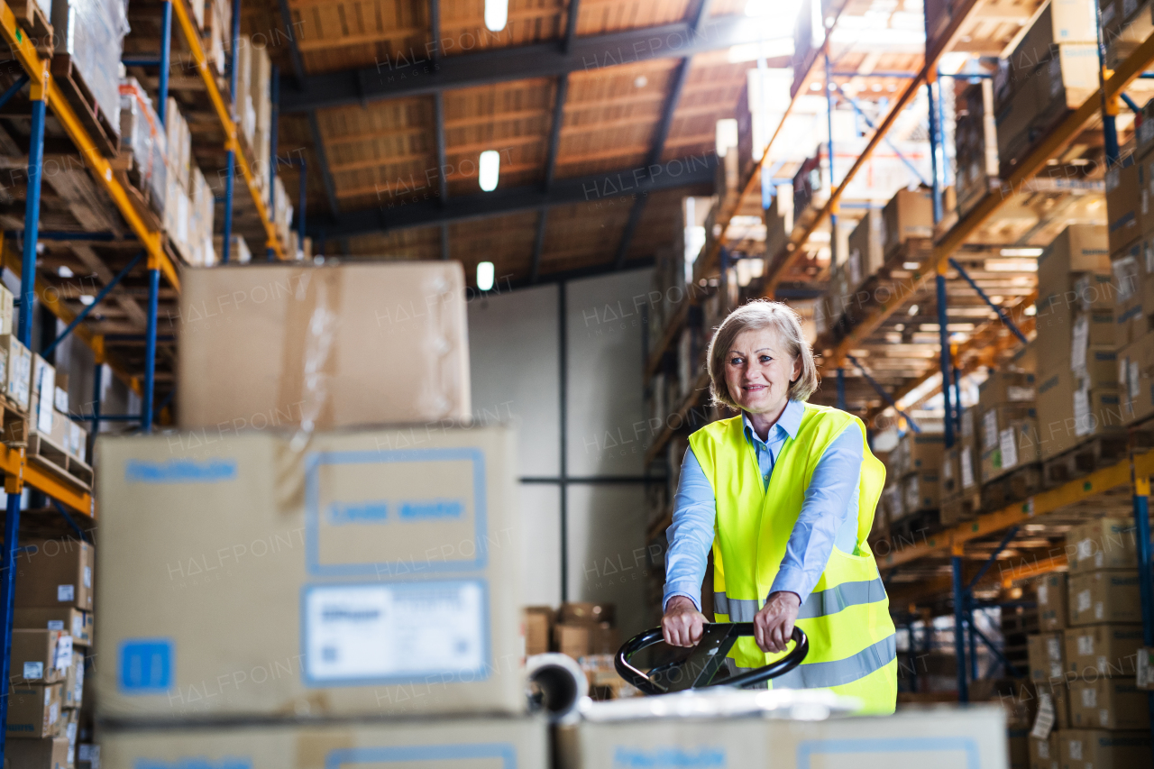 A senior woman warehouse worker or supervisor pulling a pallet truck with boxes.