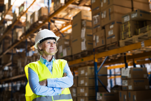 Portrait of a senior woman manager or supervisor with white helmet standing in a warehouse, arms crossed. Copy space.