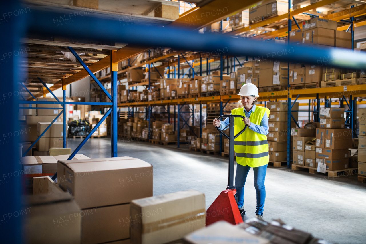 A senior woman warehouse worker or supervisor pulling a pallet truck with boxes.