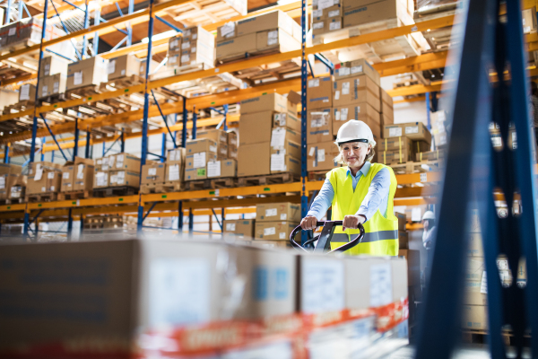 A senior woman warehouse worker or supervisor pulling a pallet truck with boxes.