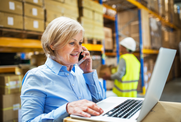 Senior woman manager with laptop and smartphone and man worker working in a warehouse.