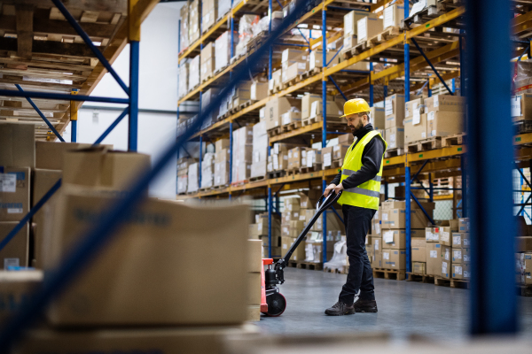 Young male warehouse worker pulling a pallet truck with boxes.