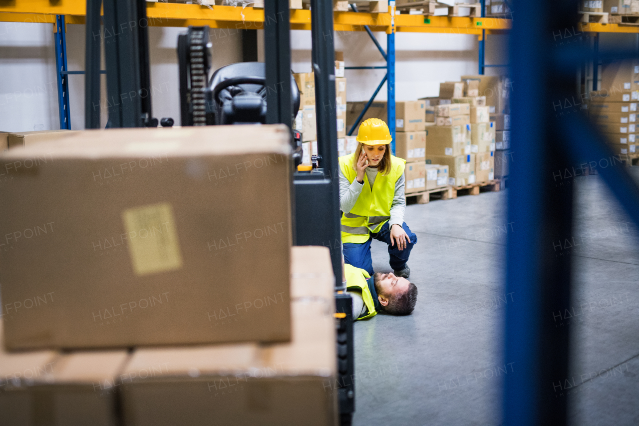 An accident in a warehouse. Woman with smartphone and her colleague lying on the floor next to a forklift.