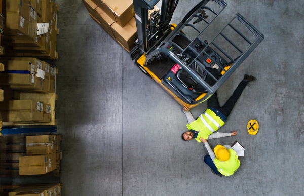 An accident in a warehouse. Woman with smartphone and her colleague lying on the floor next to a forklift. Aerial view.