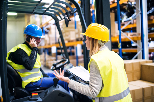 Young warehouse workers working together. Man with a smartphone, making a phone call and woman holding notes, checking something.