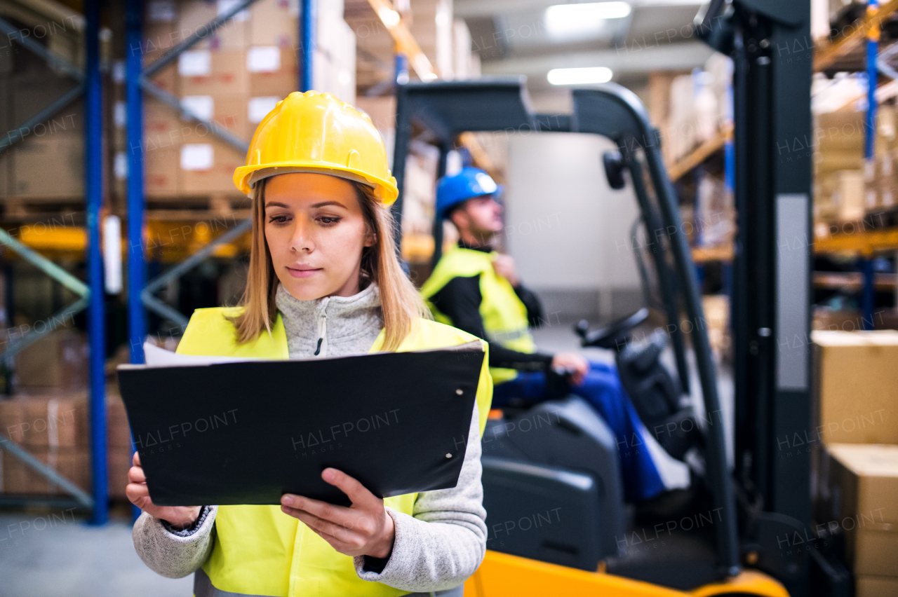 Young warehouse workers working together. Man sitting in a forklift and woman holding notes, checking something.