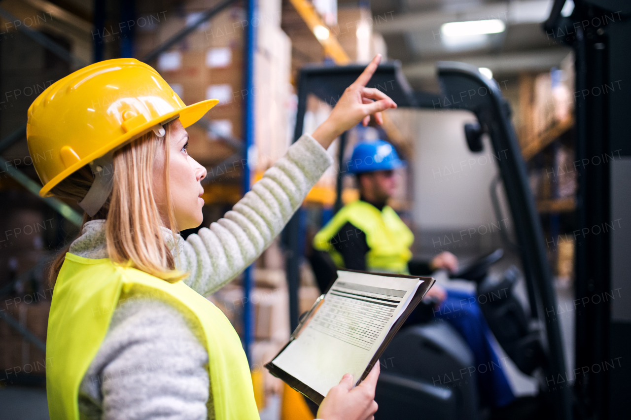 Young warehouse workers working together. Man sitting in a forklift and woman holding notes, checking something.