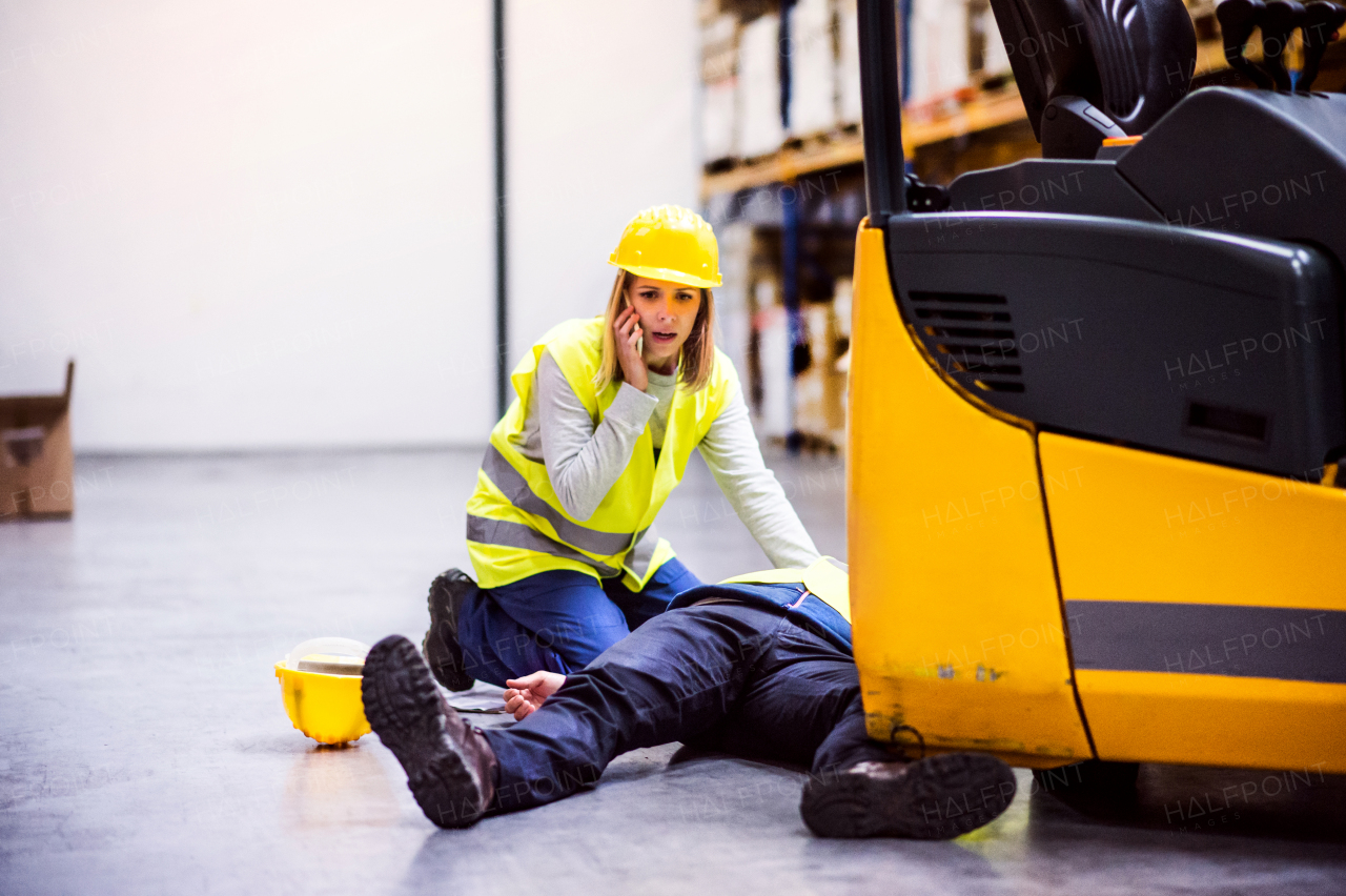 An accident in a warehouse. Woman with smartphone and her colleague lying on the floor next to a forklift.