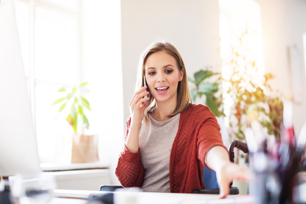 Businesswoman in wheelchair with smartphone sitting at the desk, making a phone call.