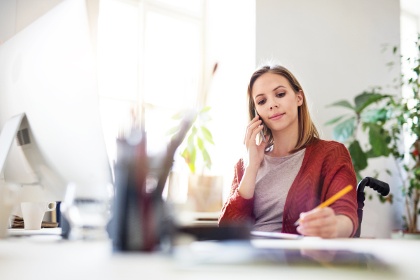 Businesswoman in wheelchair with smartphone sitting at the desk, making a phone call.