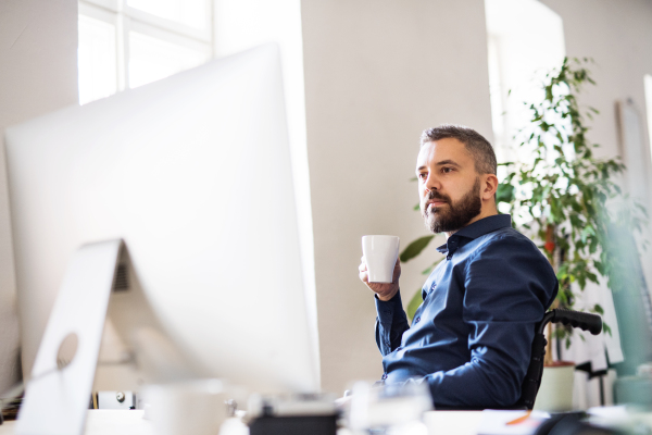 Businessman in wheelchair in the office, holding a cup of coffee.