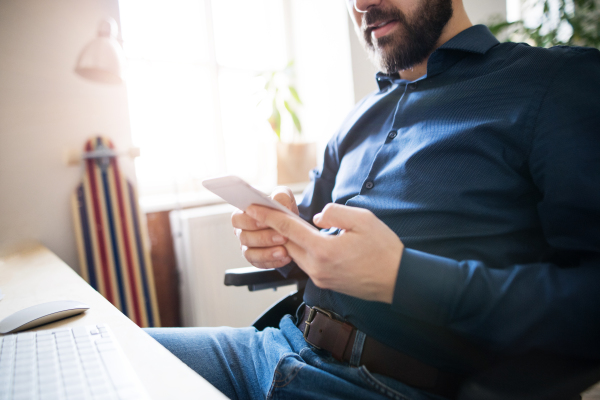 Unrecognizable businessman in wheelchair with smartphone at the desk in the office, text messaging.