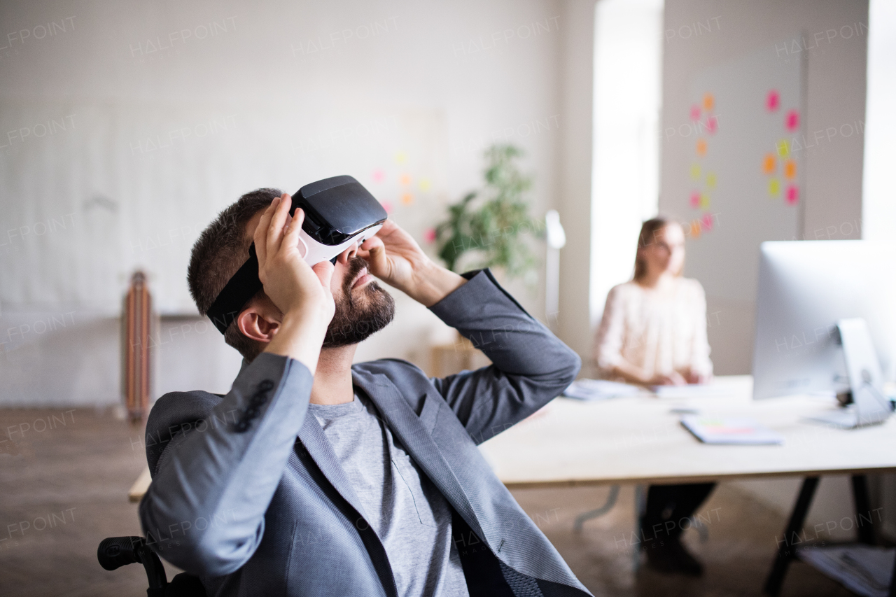 Two business people with wheelchair in the office working together. A man wearing VR goggles.