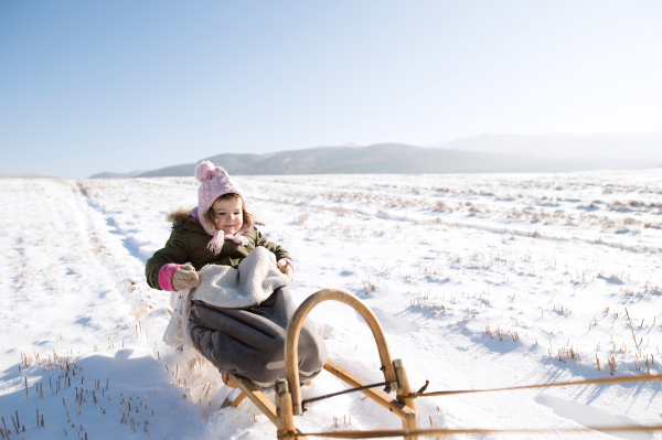 Cute little girl outside in winter nature on sunny day, sitting on sledge, being pulled by someone.