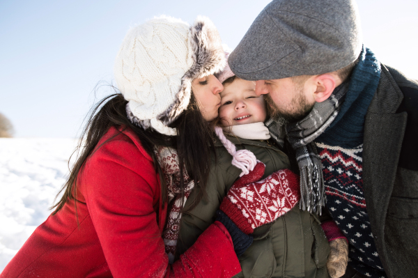 Father and mother having fun with their daughter, playing in the snow. Sunny white winter nature. Parents kissing their little daughter.