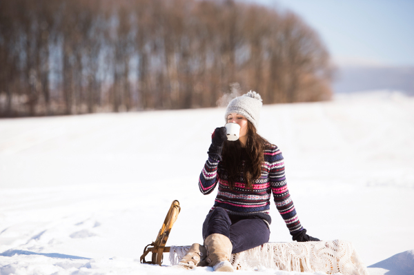 Beautiful young woman in knitted hat and gloves holding a cup of coffee outside in winter nature. Woman sitting on a sledge.