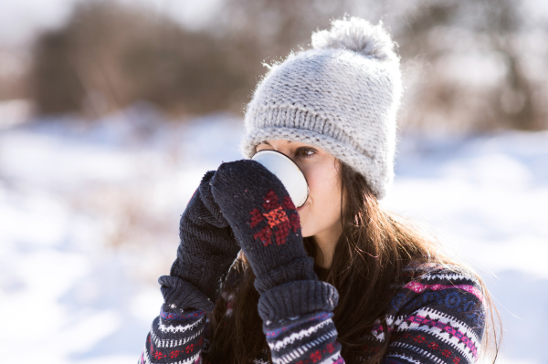 Beautiful young woman in knitted hat and gloves holding a cup of coffee outside in winter nature