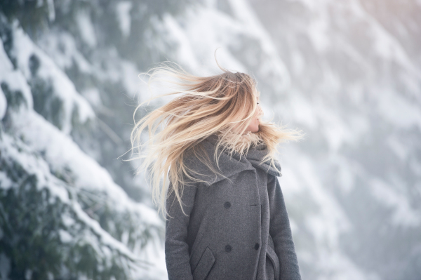 Beautiful young blonde woman in gray coat with fluttering hair covering her face on a walk in winter nature. Snowy day.