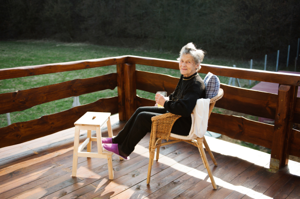 Beautiful senior woman at home, sitting on rattan chair on wooden terrace, resting with feet up on wooden step stool.