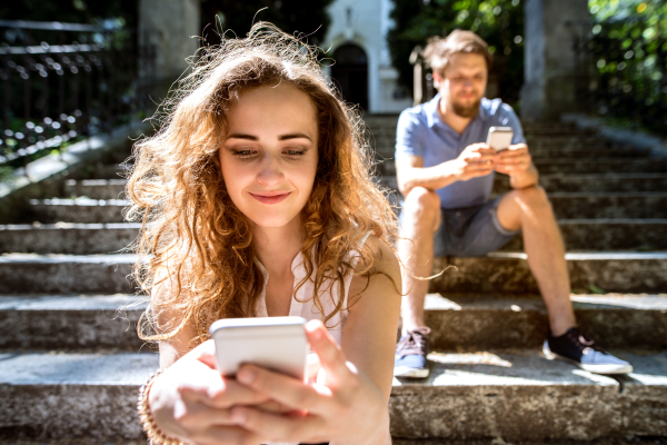 Beautiful young couple with smart phones sitting on stairs in town, texting. Sunny spring day.