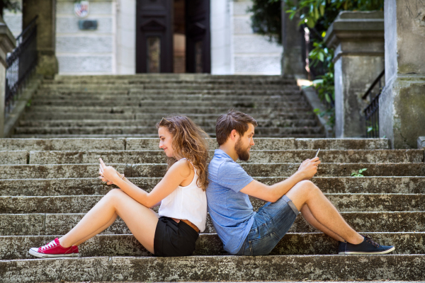 Beautiful young couple with smart phones sitting on stairs in town, texting. Sunny spring day.