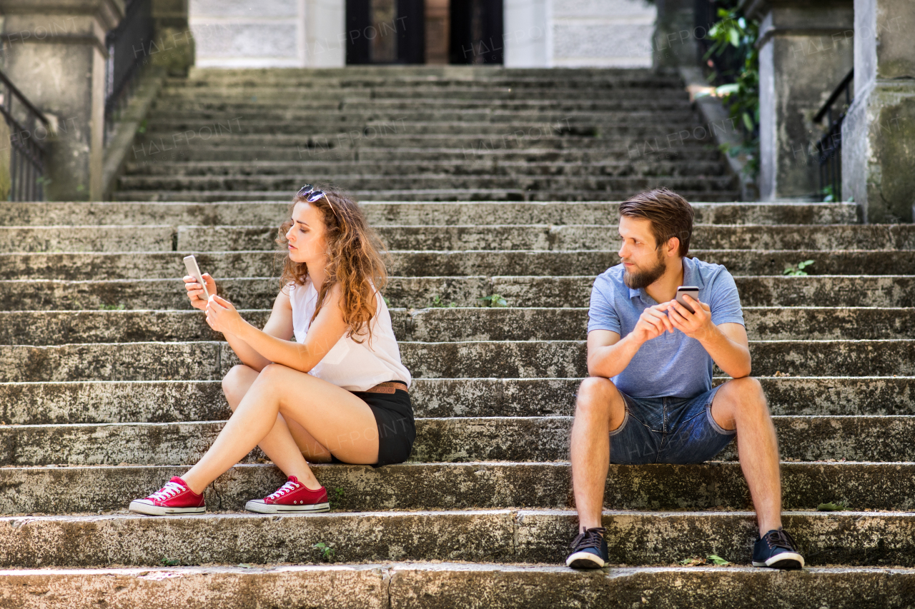 Beautiful young couple with smart phones sitting on stairs in town, texting. Sunny spring day.