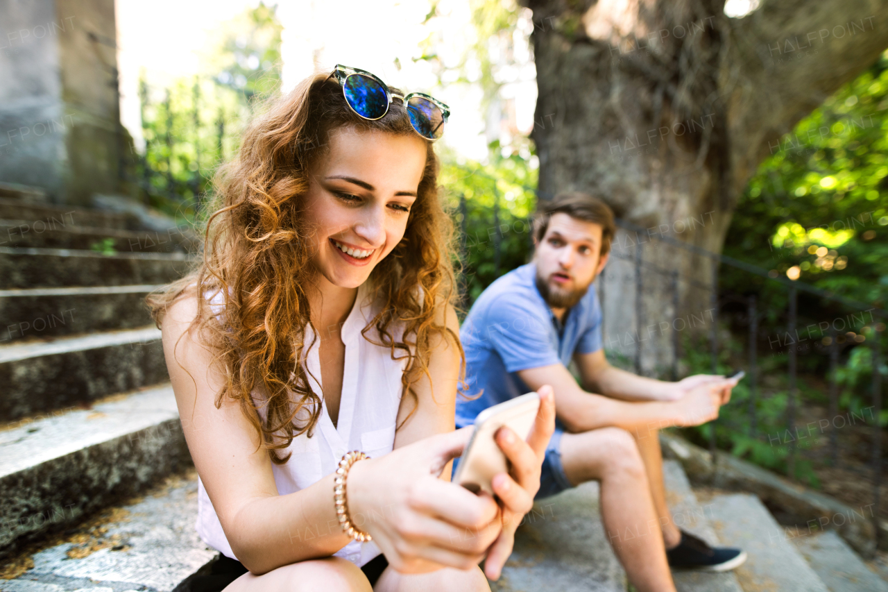 Beautiful young couple with smart phones sitting on stairs in old town, watching or reading something. Sunny spring day.