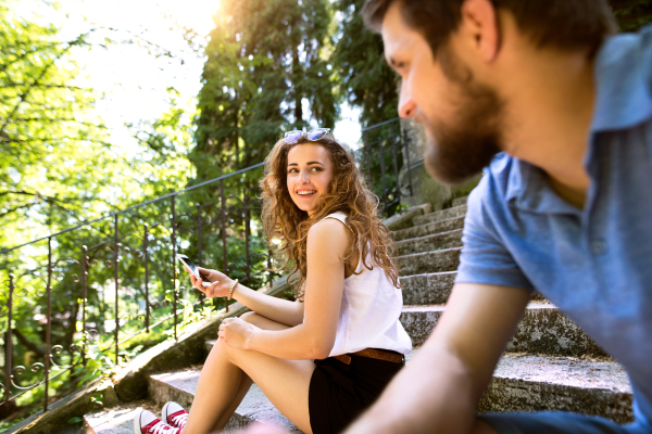 Beautiful young couple with smart phone sitting on stairs in old town, watching or reading something. Sunny spring day.