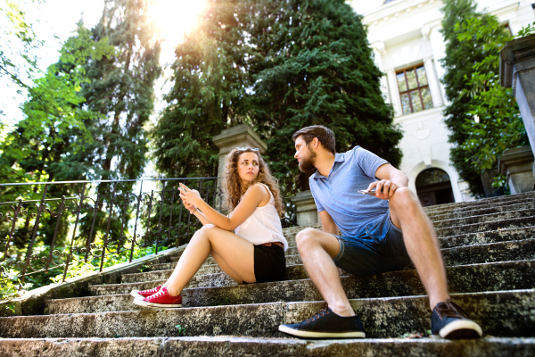 Beautiful young couple with smart phone sitting on stairs in old town, watching or reading something. Sunny spring day.