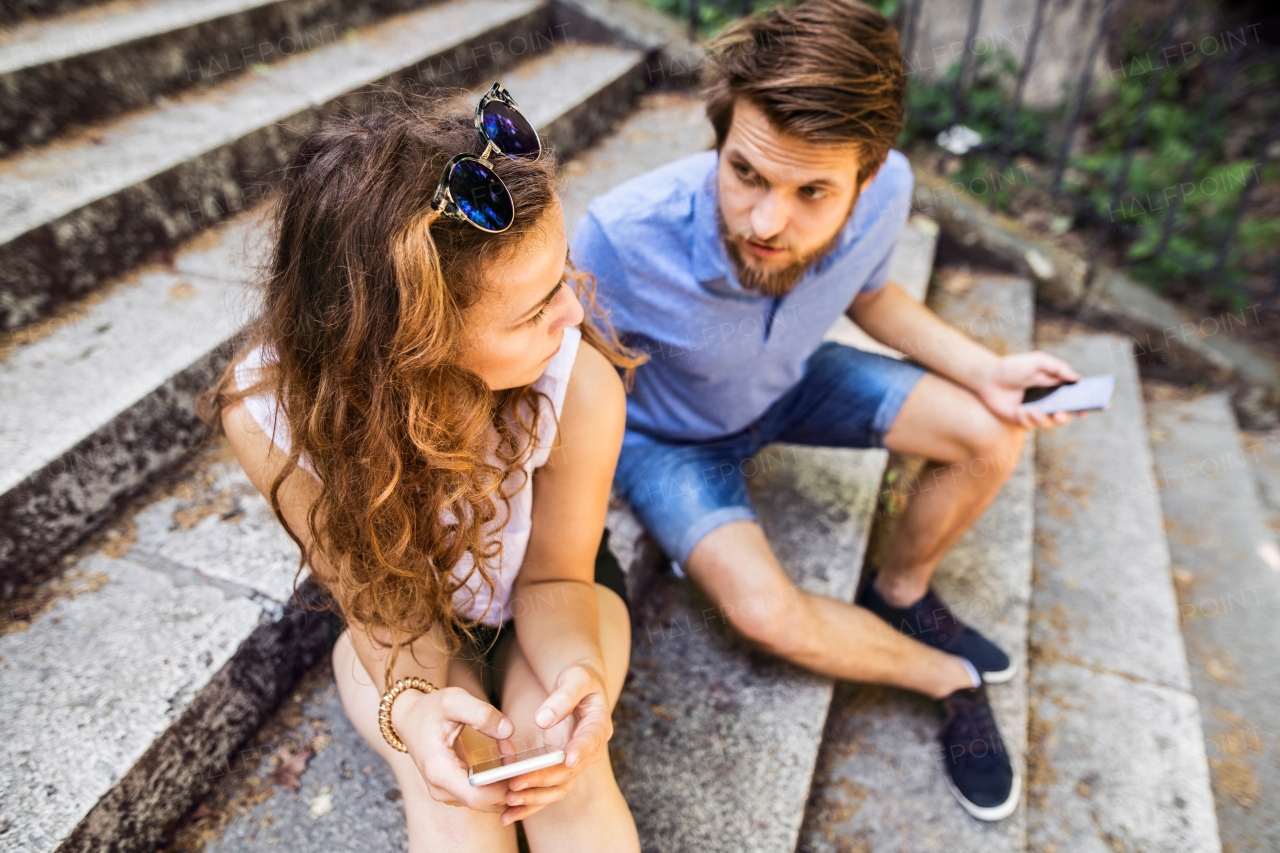 Beautiful young couple with smart phones sitting on stairs in town, texting. Sunny spring day.