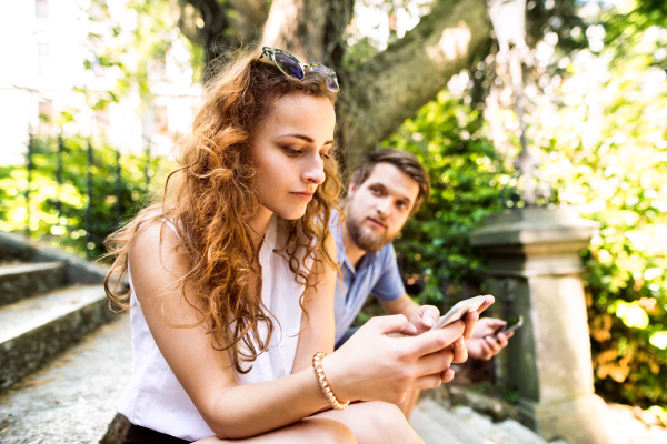 Beautiful young couple with smart phones sitting on stairs in town, texting. Sunny spring day.