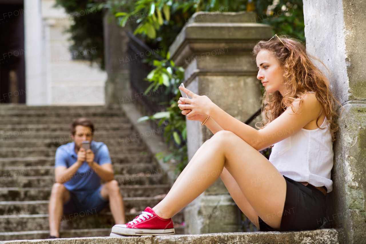 Beautiful young couple with smart phones sitting on stairs in town, watching or reading something. Sunny spring day.