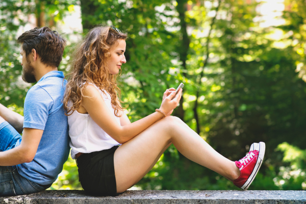 Beautiful young couple in town sitting on concrete wall, holding smart phones, watching or reading something. Sunny spring day.