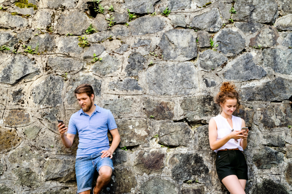 Beautiful young couple with smart phones standing against stone wall in old town, watching or reading something. Sunny spring day.