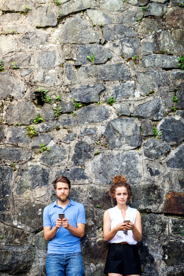 Beautiful young couple with smart phones standing against stone wall in old town, watching or reading something. Sunny spring day.