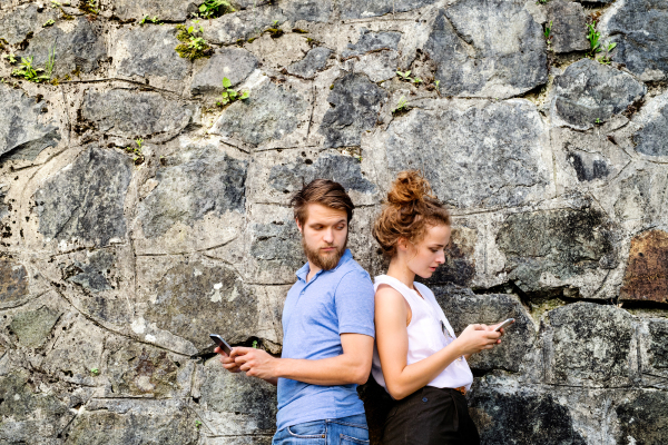 Beautiful young couple with smart phones standing against stone wall in old town, watching or reading something. Sunny spring day.