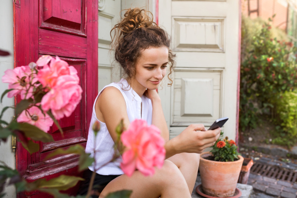 Young woman with a smartphone sitting at the doorstep, reading something. Sunny spring day.