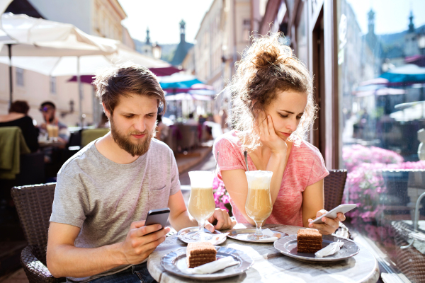 Beautiful young couple with smart phones sitting in cafe, watching or reading something. Sunny spring day.