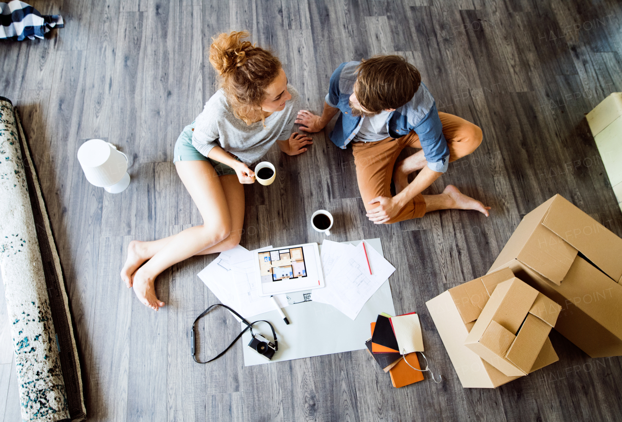 Young married couple moving in new house, sitting on the floor near cardboard boxes, drinking coffee.