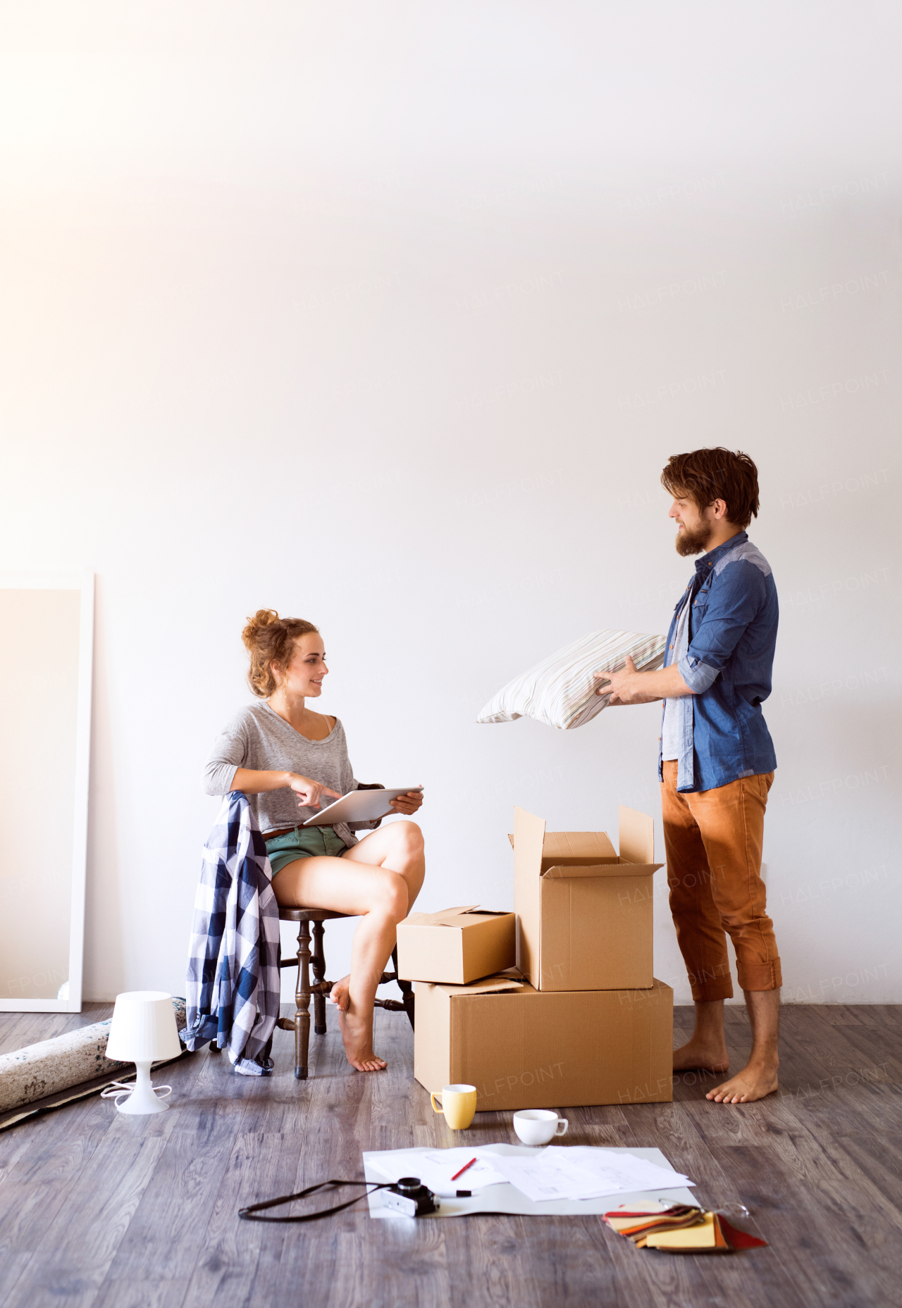 Young married couple moving in new house, woman with tablet sitting on chair, man unpacking things from a cardboard box.