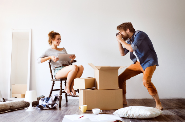 Young married couple moving in new house, woman with tablet sitting on chair, man taking pictures of her.