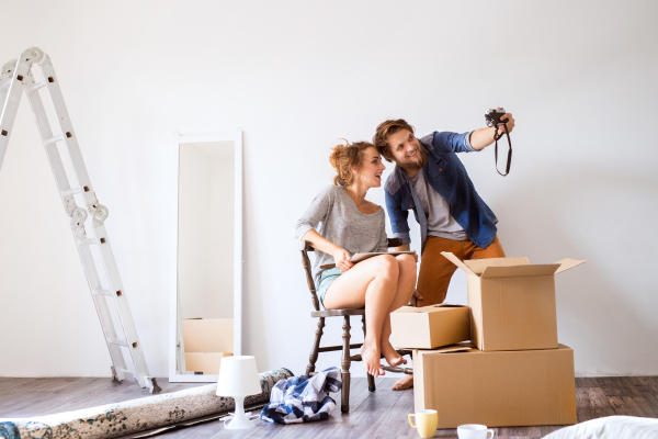 Young married couple moving in a new house, woman with tablet sitting on chair, man taking selfie of them.