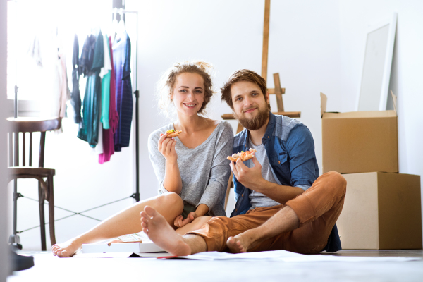 Young married couple moving in new house, sitting on the floor near cardboard boxes, eating pizza.