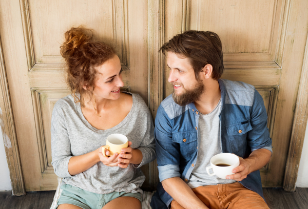 Young married couple moving in new house, sitting on the floor agaings old wooden door, drinking coffee, resting