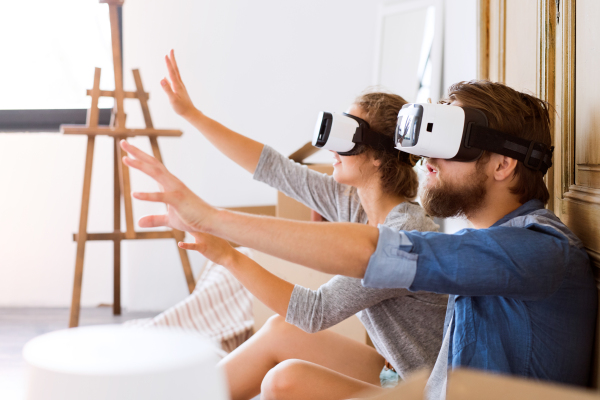 Young married couple moving in new house, sitting on the floor, wearing virtual reality goggles.